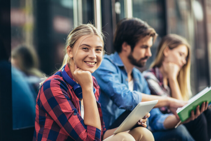 Junge Frau mit Tablet in der Hand l&auml;chelt in die Kamera, im Hintergrund unscharf sitzen ein weiterer Mann und eine Frau mit Unterlagen in der Hand
