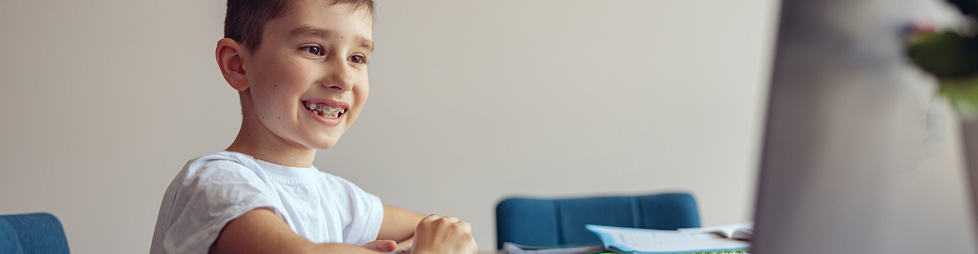 Kleiner Bub mit Zahnspange sitz an einem Tisch mit vielen Schulb&uuml;cher, hat Stift in der Hand und schaut l&auml;chelnd in einen Computerbildschirm