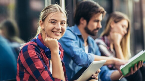 Junge Frau mit Tablet in der Hand l&auml;chelt in die Kamera, im Hintergrund unscharf sitzen ein weiterer Mann und eine Frau mit Unterlagen in der Hand