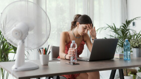 Junge Frau in leichtem Sommerkleid sitzt am Schreibtisch und wischt sich Schweiß von der Stirn. Vor ihr ein Ventilator und zwei Wasserflaschen.