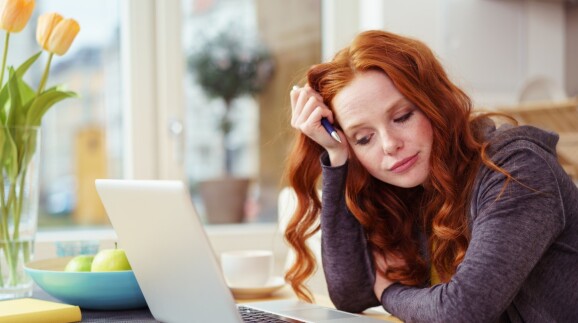 Junge Frau sitzt am K&uuml;chentisch und schaut auf ihren Laptop.