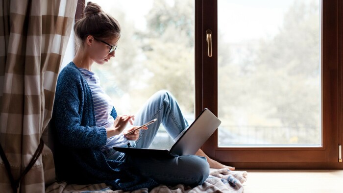 Junge Frau sitzt mit Smartphone und Laptop am Fenster