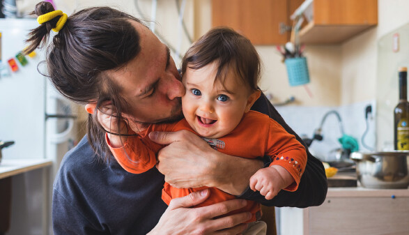 Vater sitzt mit Baby in einer chaotischen Küche und gibt ihm einen Kuss auf die Wange