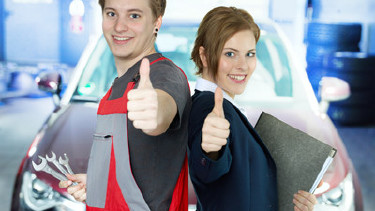 Mann in Arbeitshose und Frau im Anzug stehen vor einem Fahrzeug in einer Werkstatt und geben beide einen Daumen nach oben.